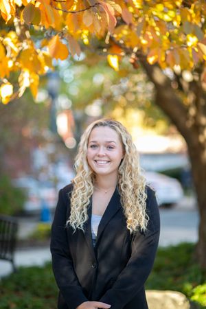 Smiling blonde woman in blazer stands under golden autumn leaves in an outdoor setting.