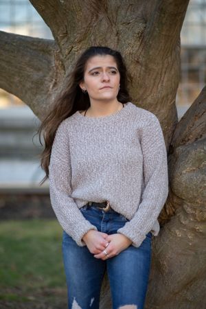 Young woman with long dark hair leans against a tree in a park wearing a sweater and jeans.