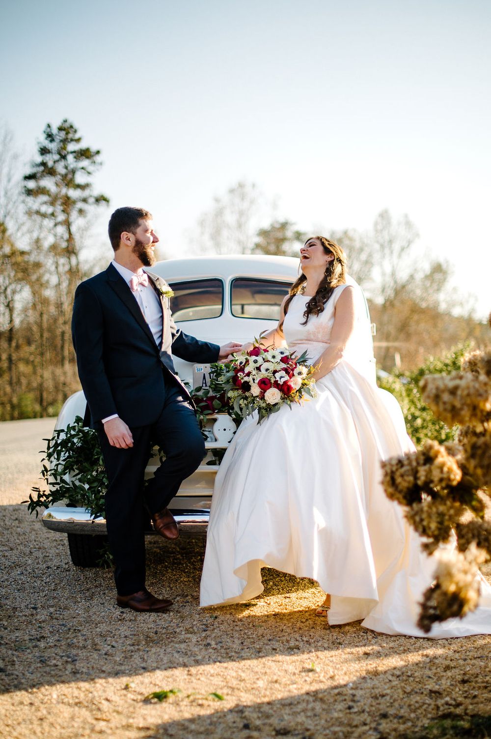 Laughing bride and groom pose beside a vintage white car holding a red and white bouquet.