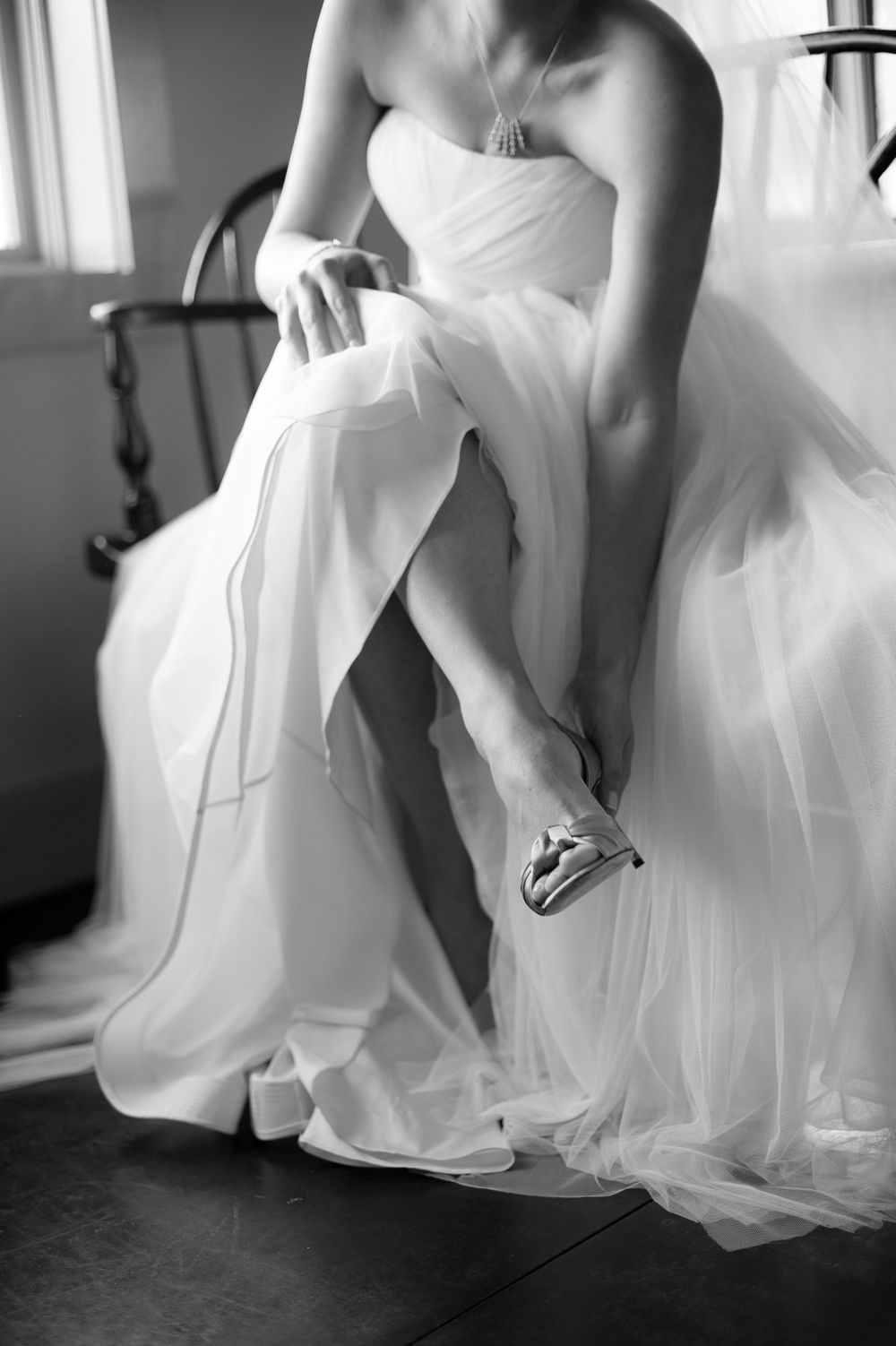 Bride in flowing white tulle gown putting on silver heels in black and white wedding preparation photo.