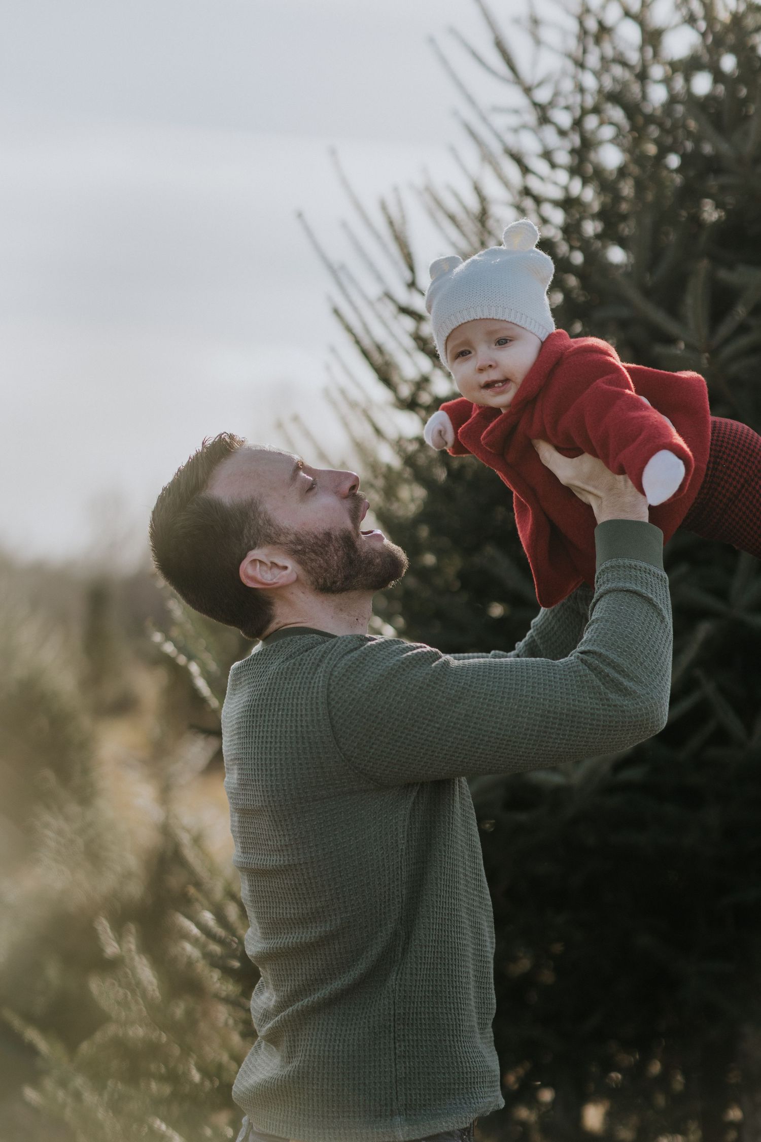 Parent in green sweater playfully lifting baby wearing red coat in the air.