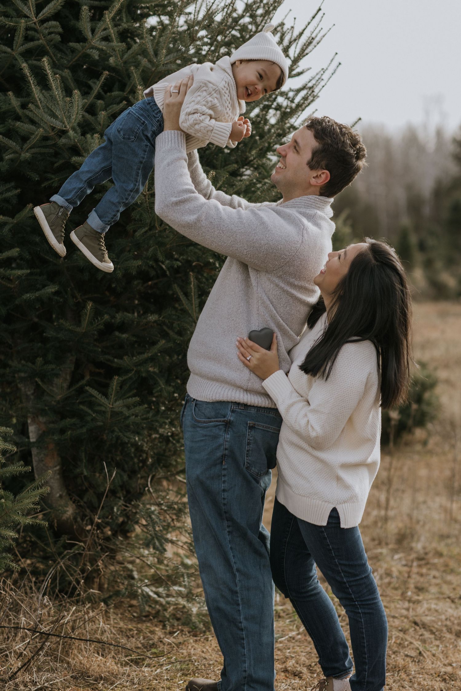 Parents lifting and playing with small child among evergreen trees.