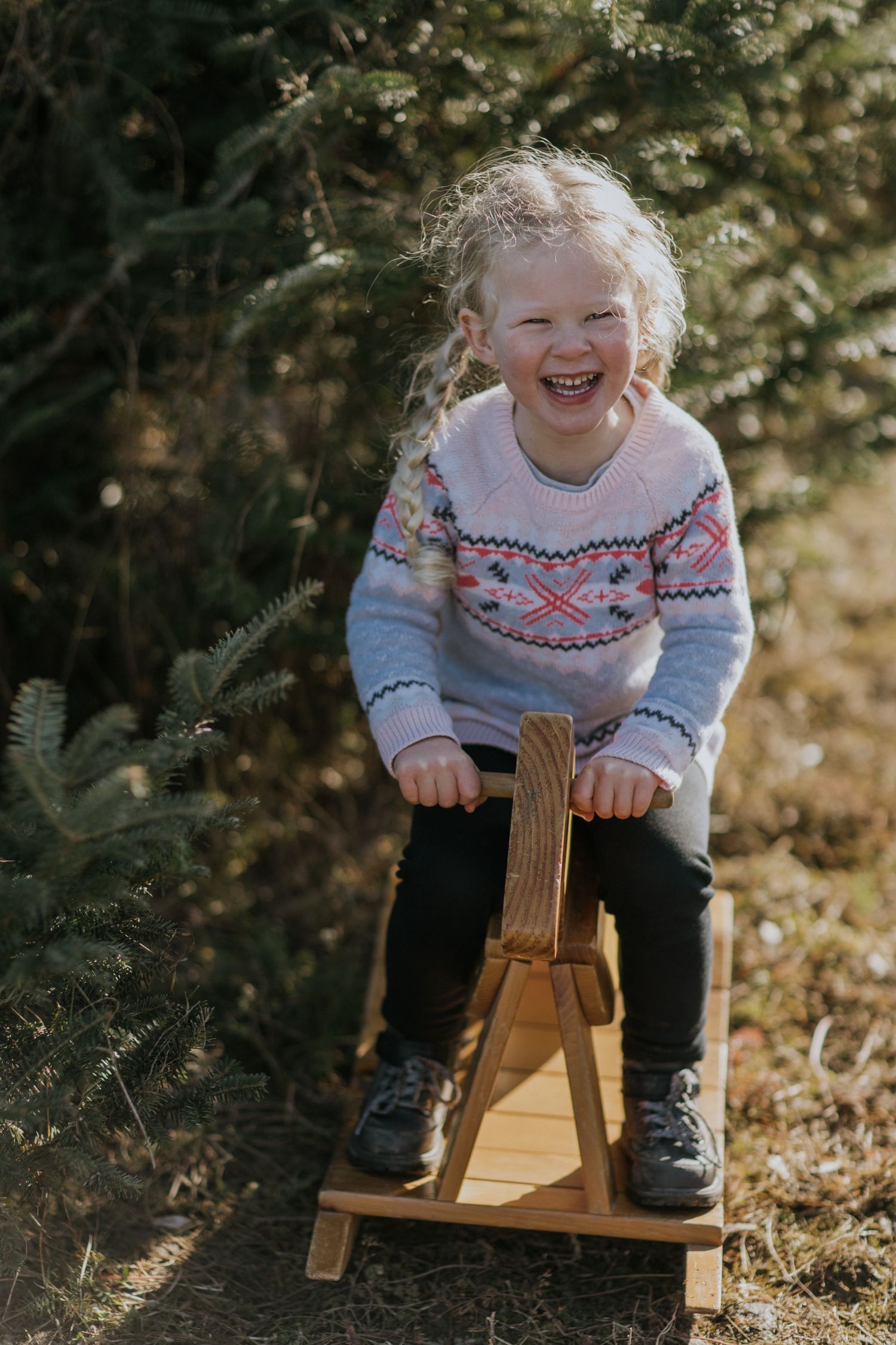 Small child wearing a festive sweater joyfully riding a wooden rocking toy outdoors.