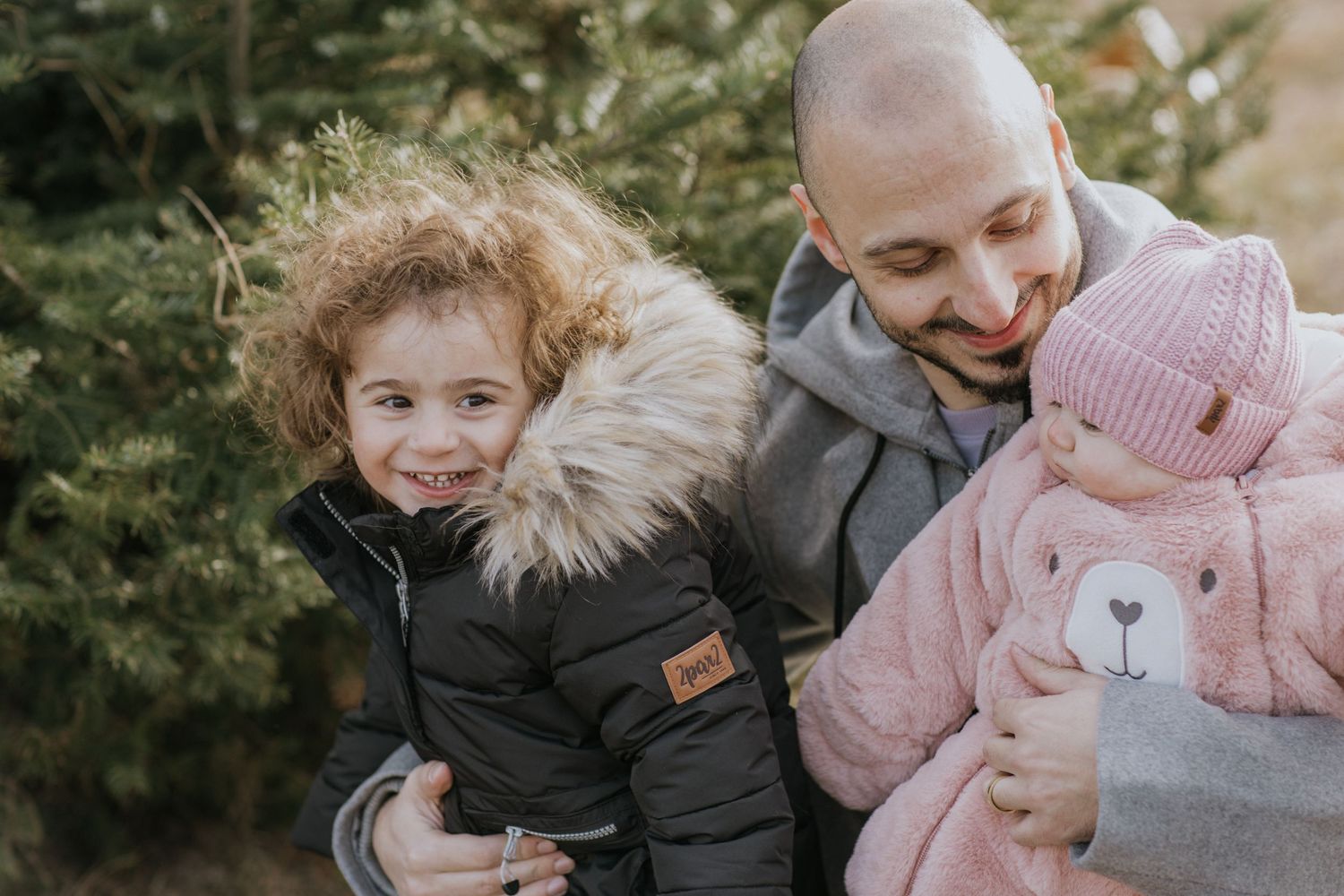 Family walking together through winter evergreen trees with small child in pink coat.