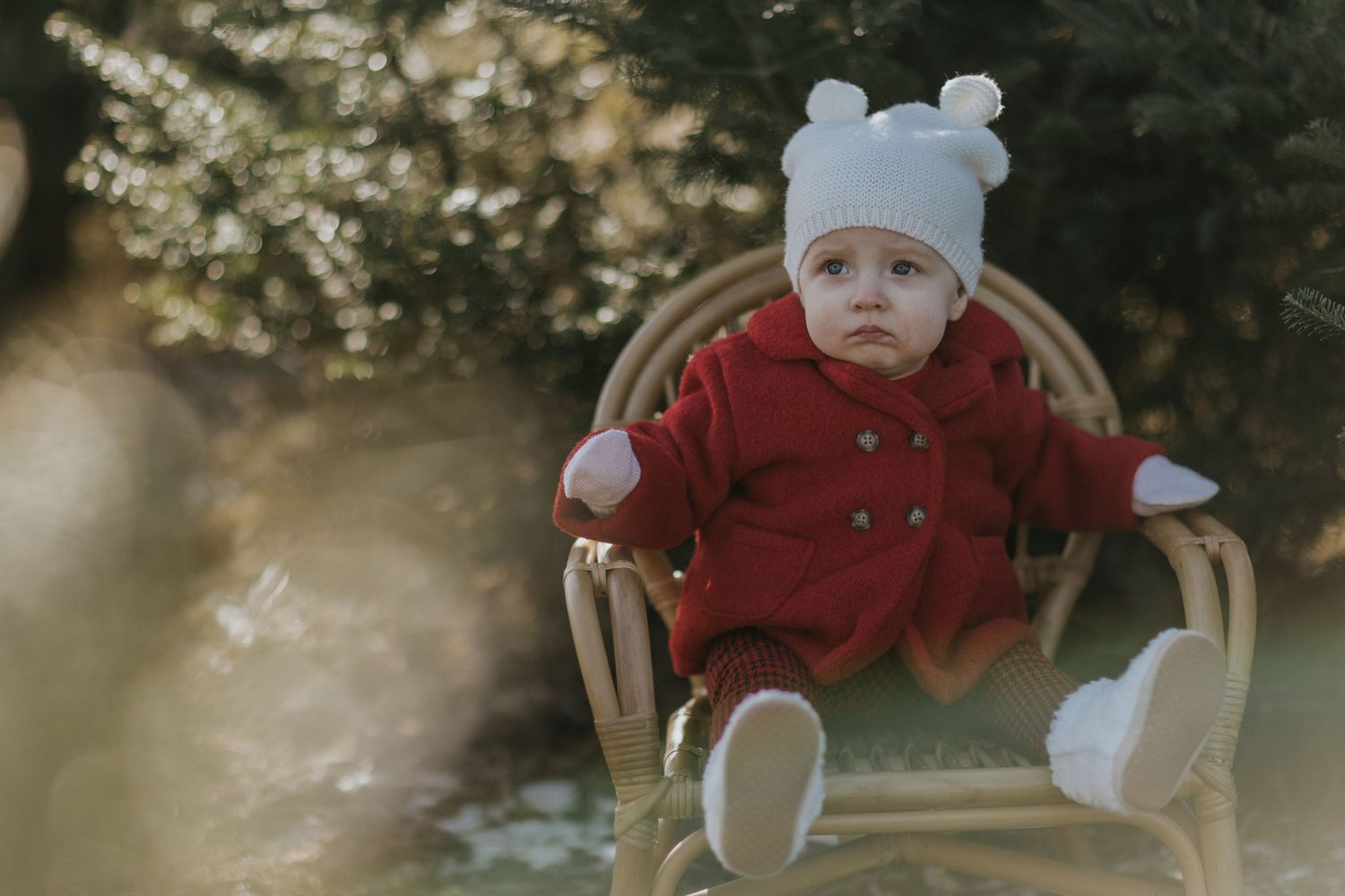 Baby wearing red coat and winter hat sitting in wooden rocking chair outdoors.