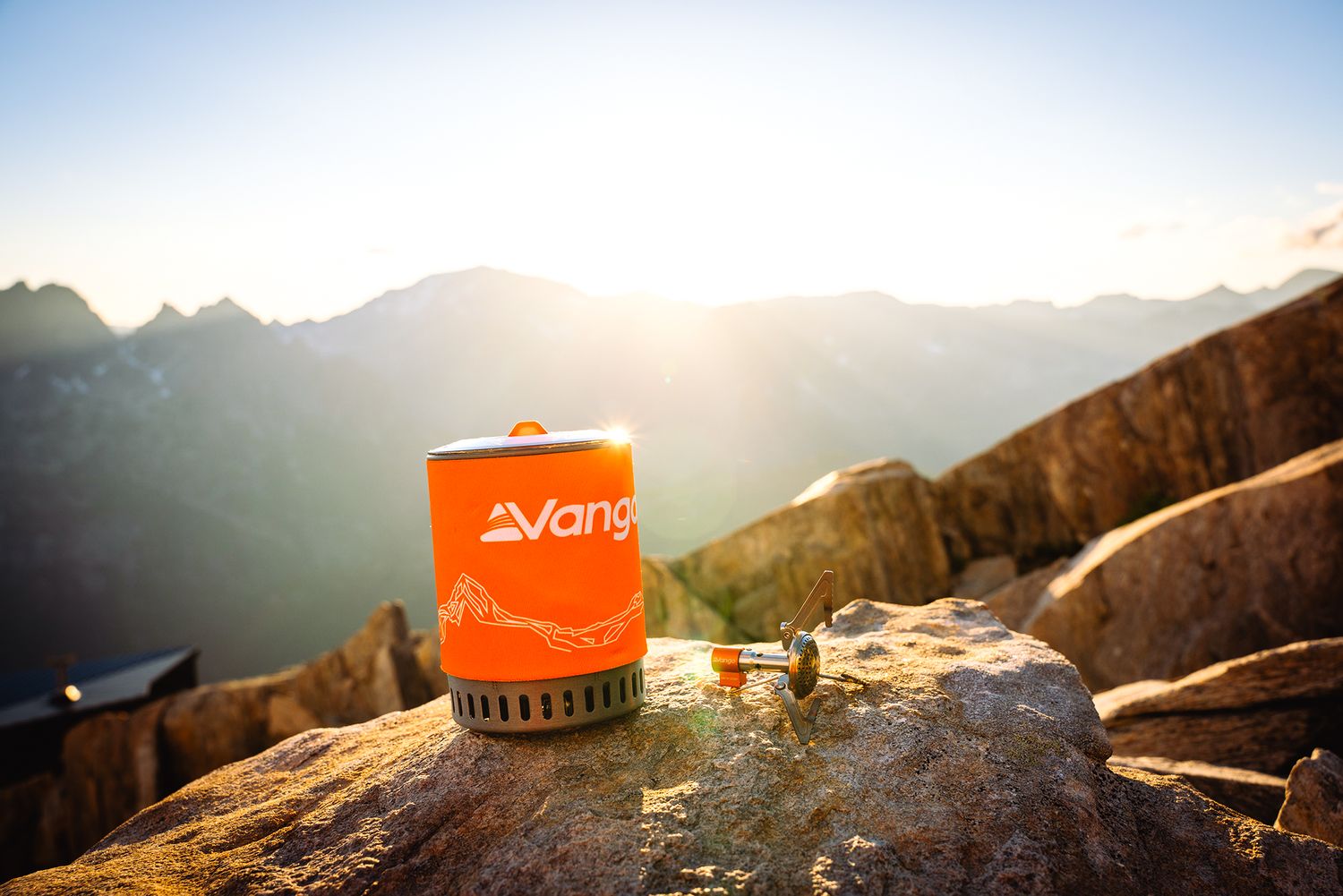 Orange camping stove perched on rocky mountain summit during golden hour with distant peaks.