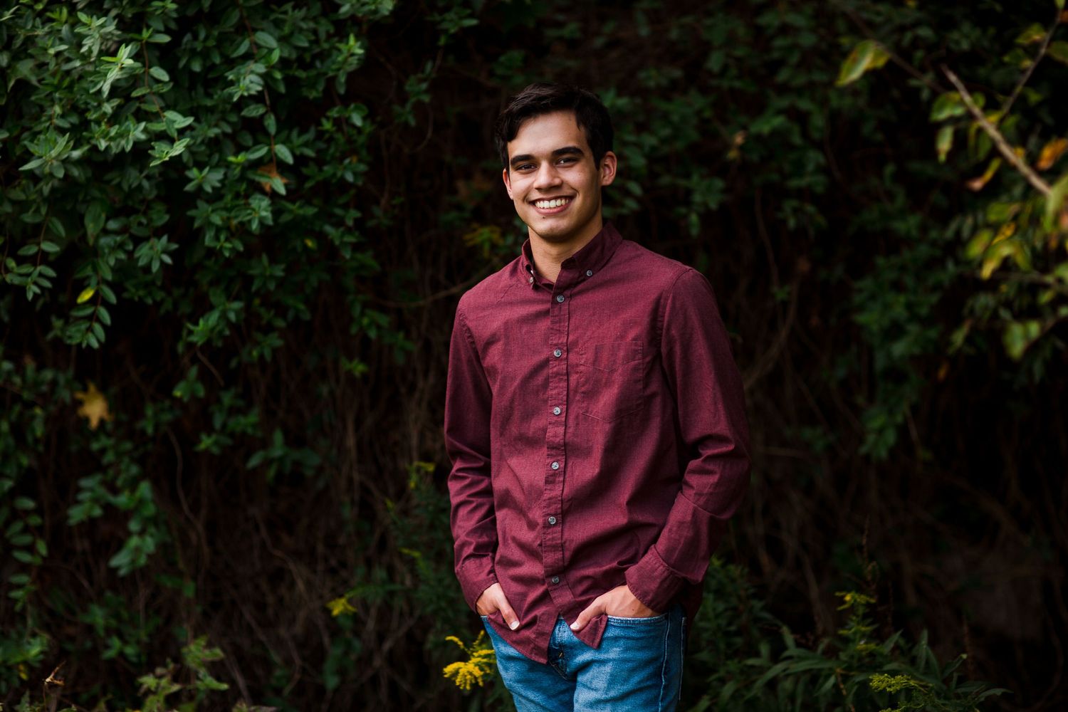 Casual portrait against green foliage background wearing burgundy button-down shirt.