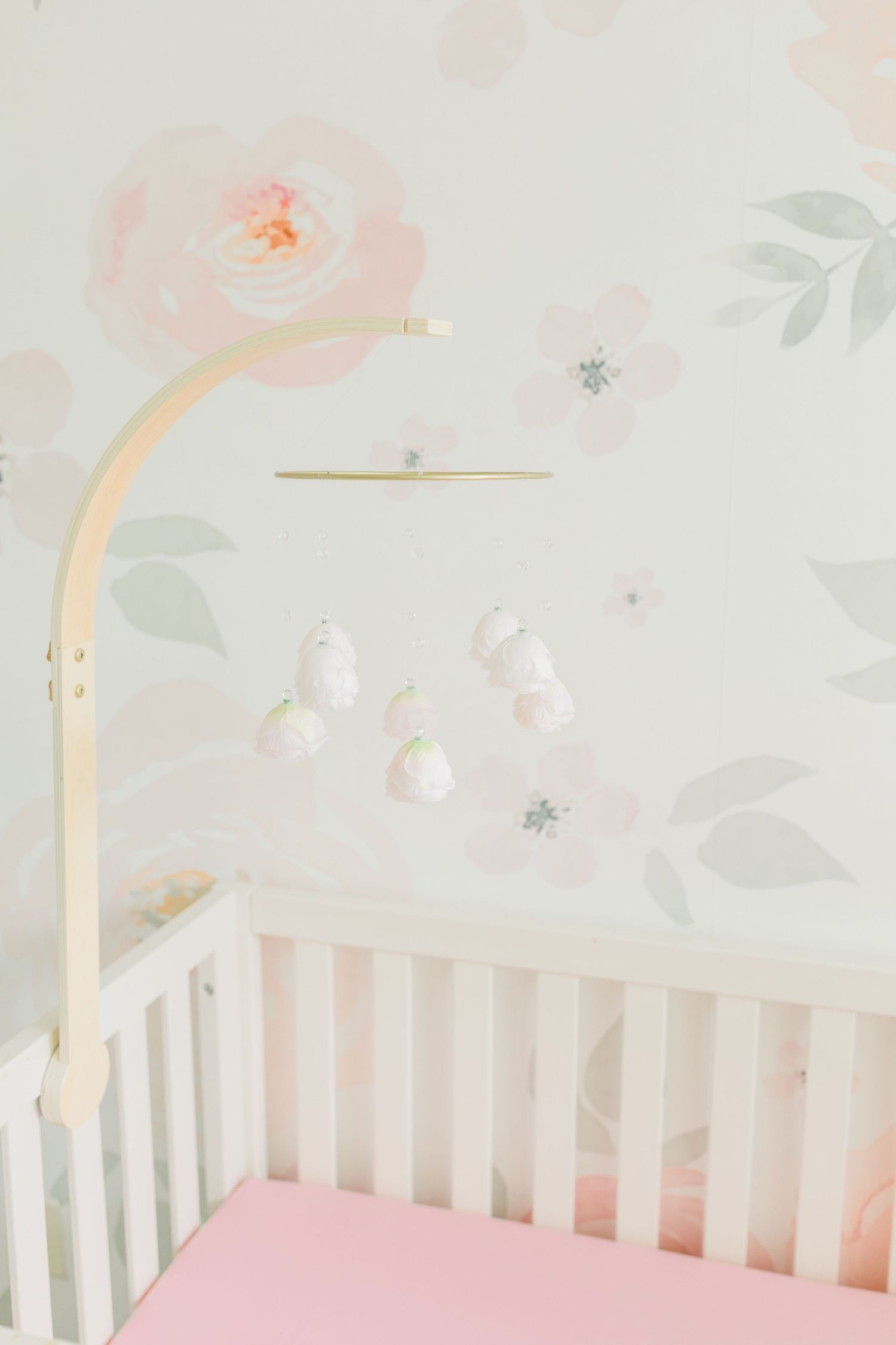 White crib with gold mobile arm against floral wallpaper in soft pink and gray.