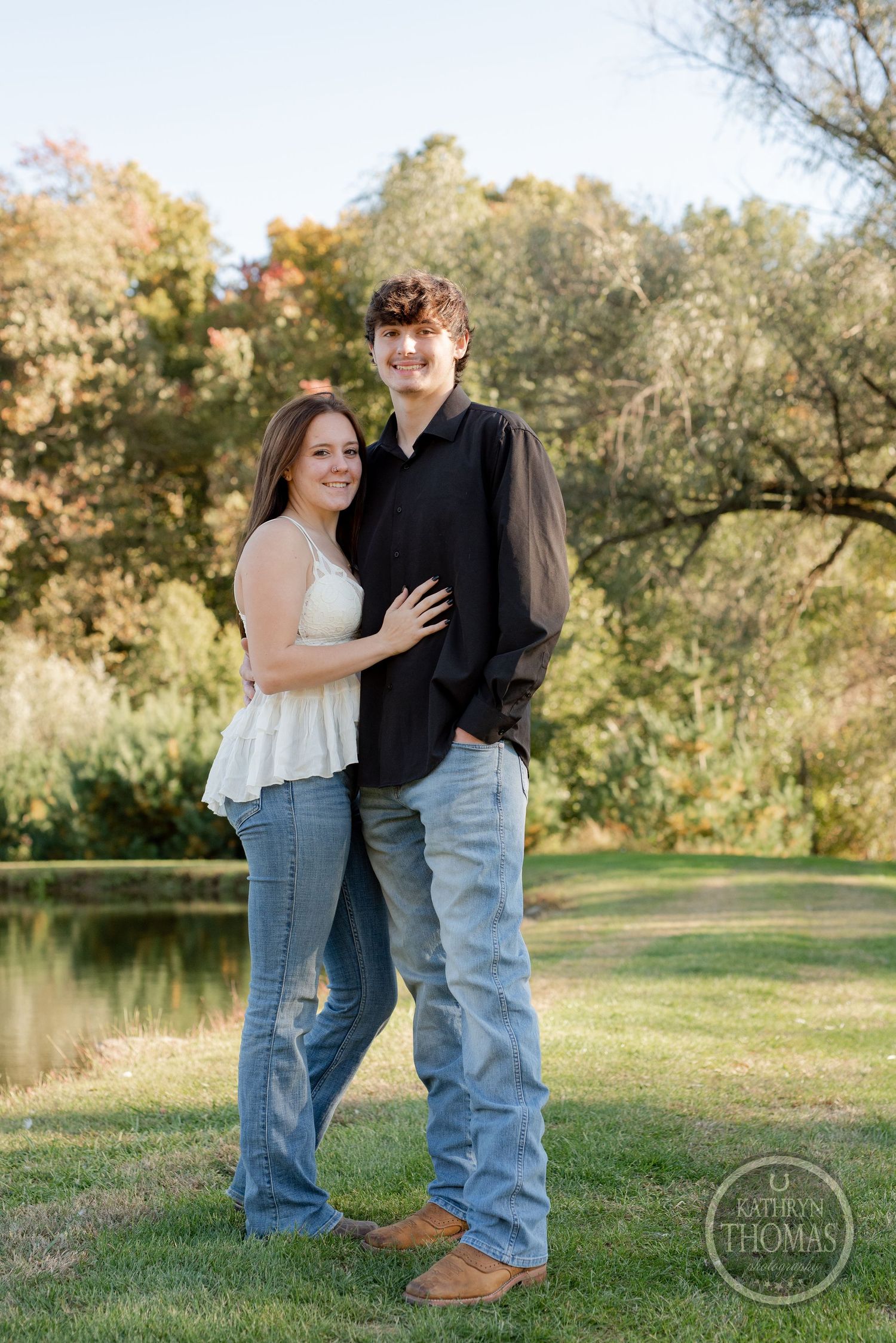 A couple poses together outdoors with a scenic pond and autumn trees in the background.