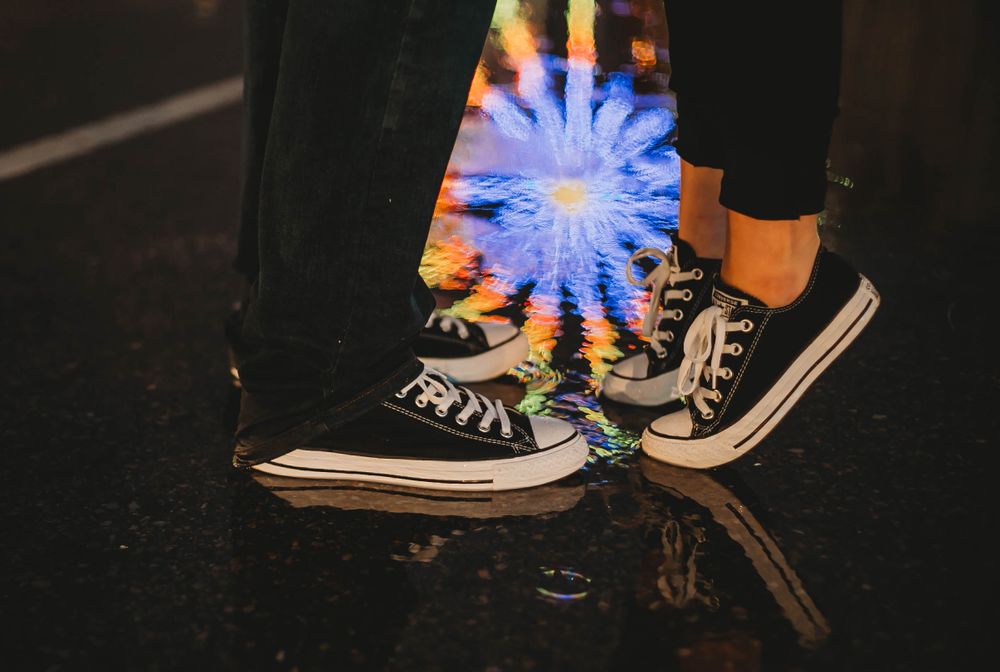 Black Converse sneakers worn with reflective ferris wheel on the pavement creating vibrant nighttime atmosphere.