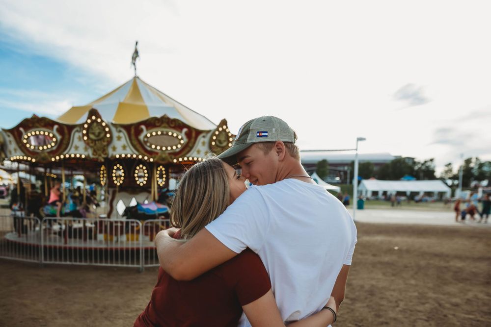 A couple embraces while standing near a vintage carousel at a fairground on a summer afternoon.