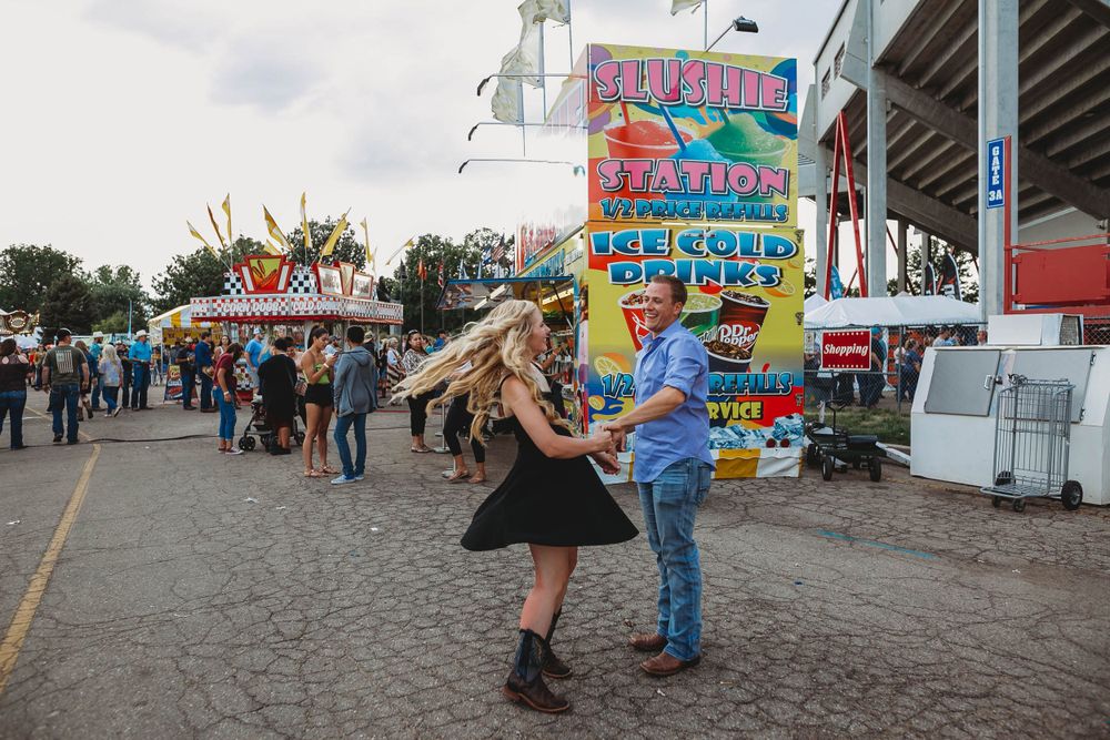 People dance near colorful carnival food stands and rides at a state fair.