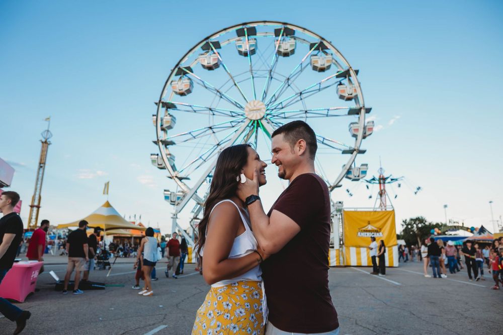 A couple embraces in front of a Ferris wheel at a carnival during golden hour with festival tents in the background.