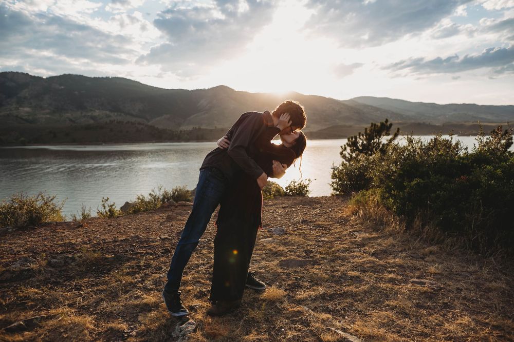 Two silhouetted figures embrace on a hilltop overlooking a scenic lake and mountains during golden hour.