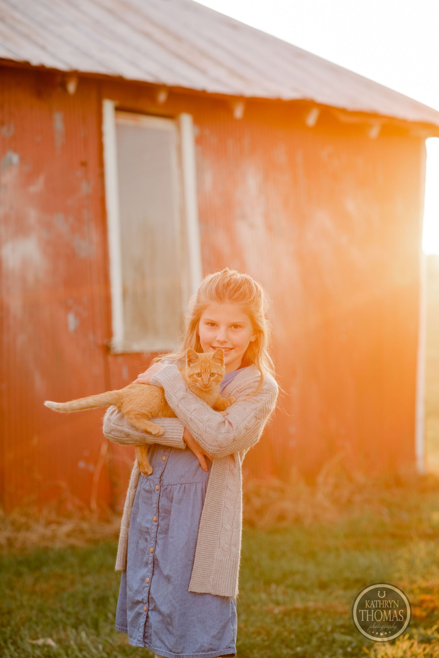 A person holds an orange cat while standing by a rustic building at sunset.
