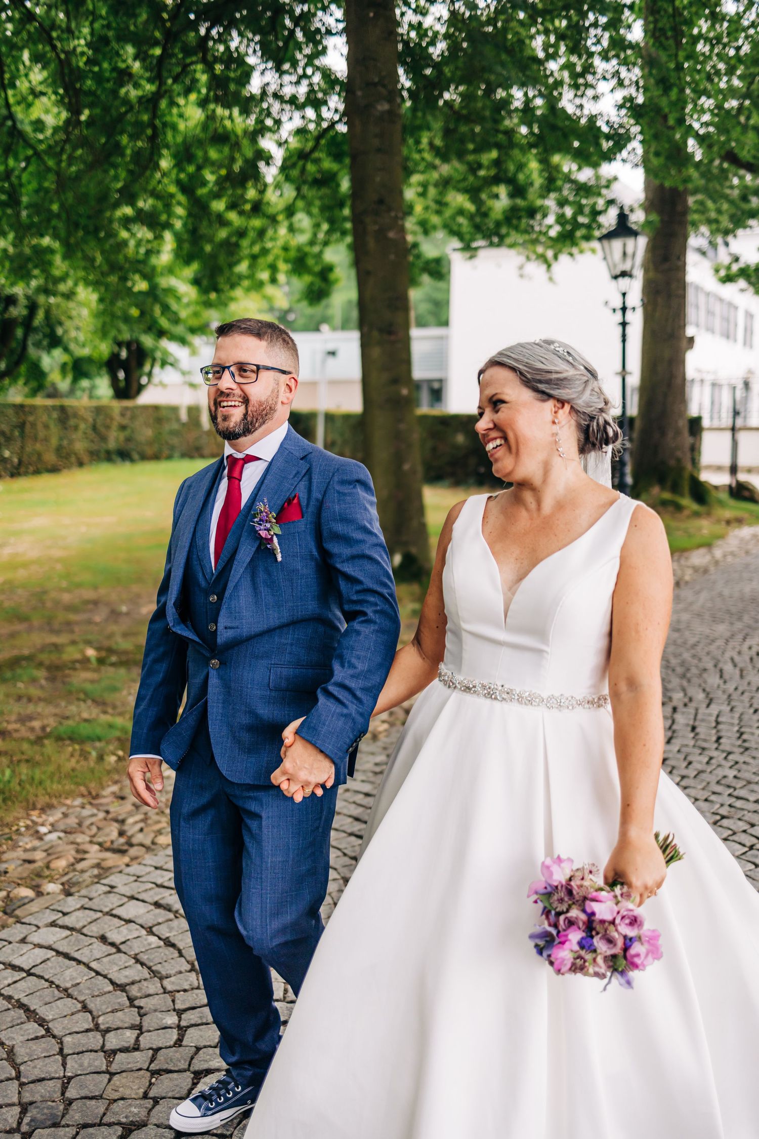 A joyful couple in wedding attire walking hand in hand on a cobblestone path surrounded by trees.