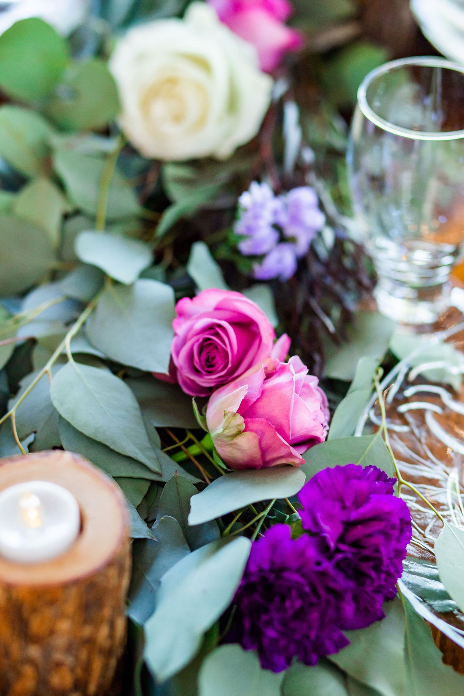 Table centerpiece with pink roses, purple flowers, and eucalyptus leaves next to wine glasses and candles.