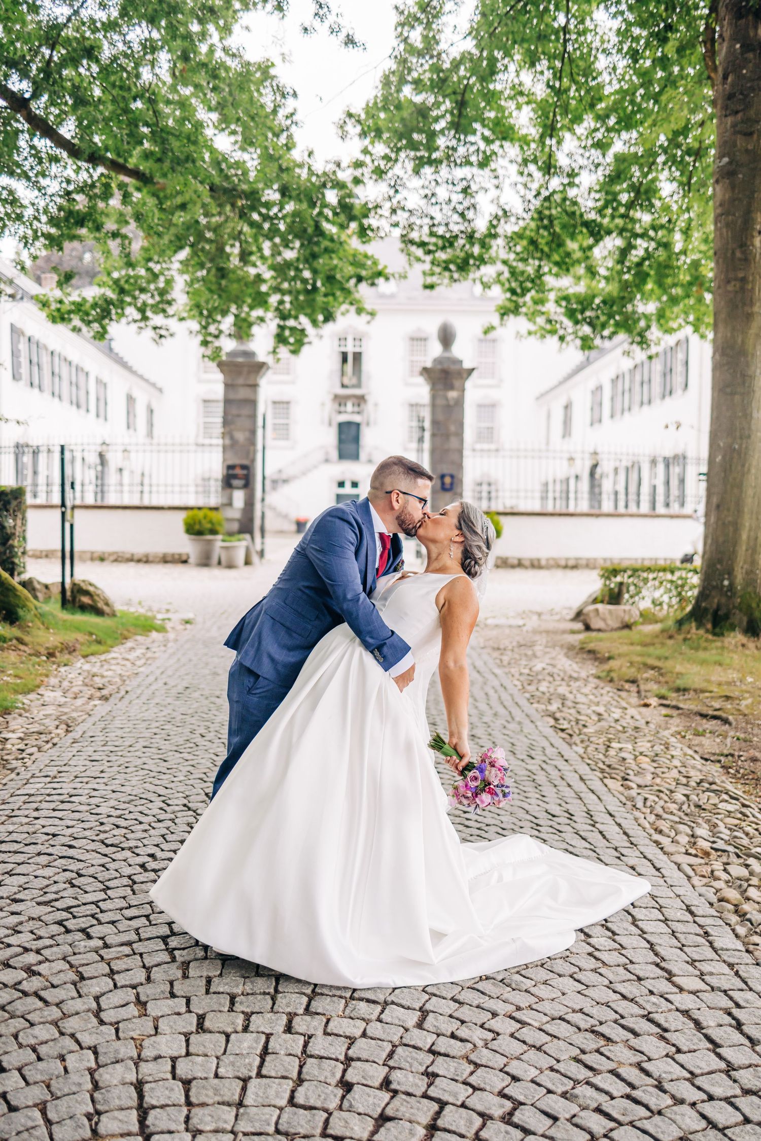 A romantic wedding couple sharing a kiss on cobblestone path with elegant manor house backdrop.