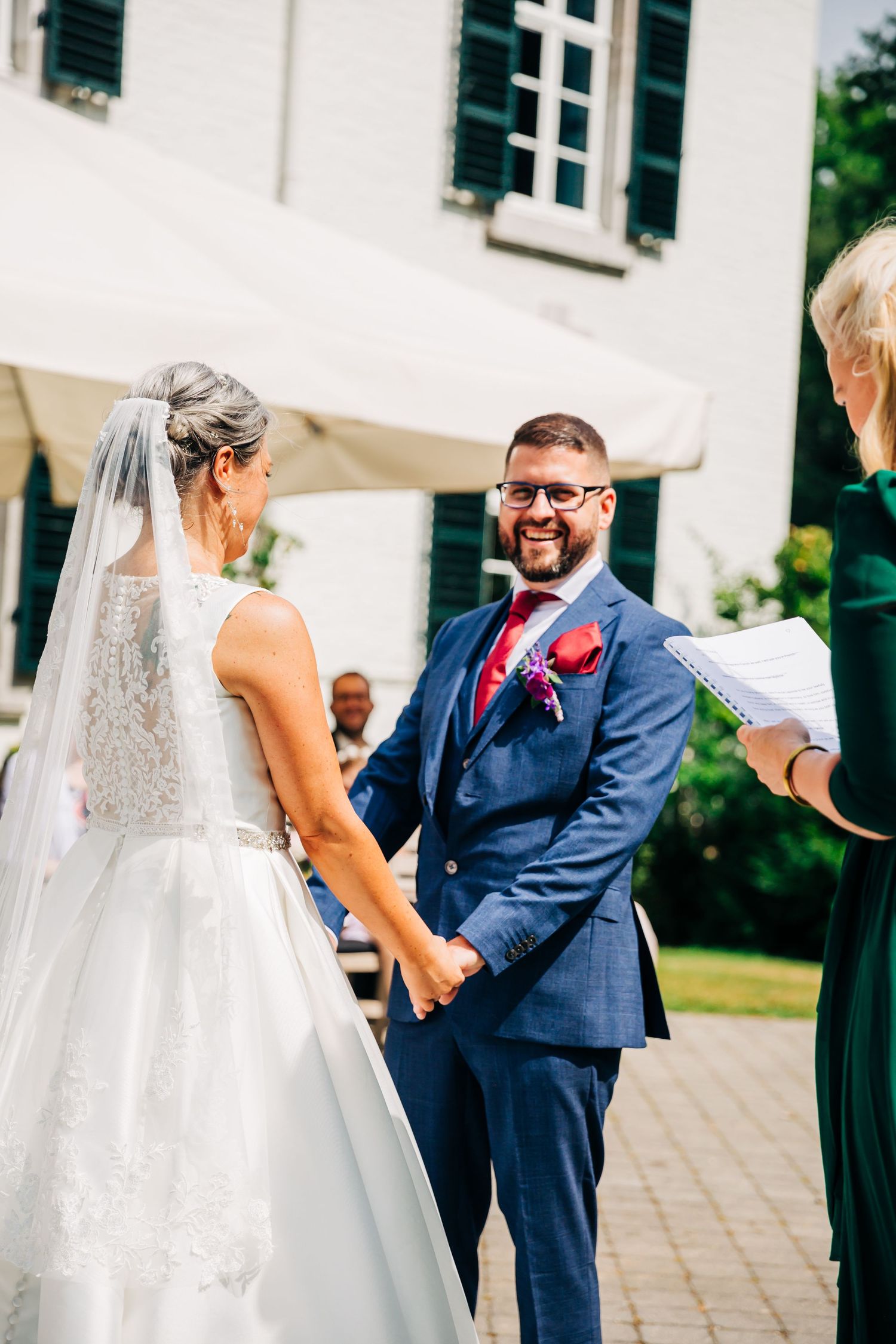 A wedding ceremony moment outdoors with white building and green shutters in background.