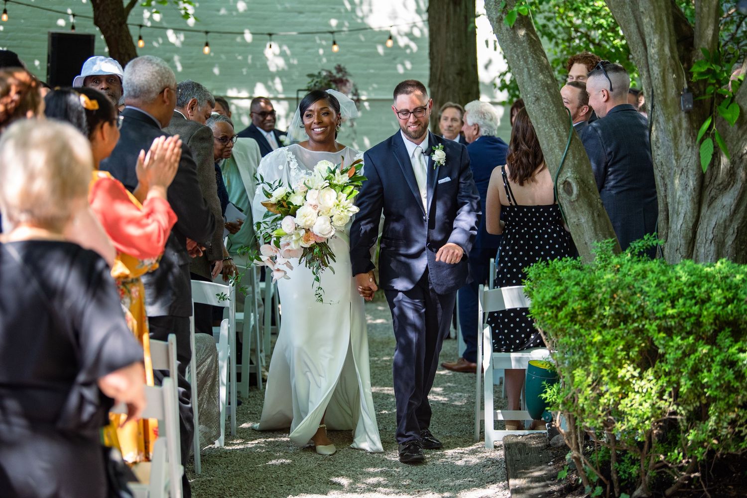 Wedding guests gather under trees during an outdoor garden ceremony celebration.