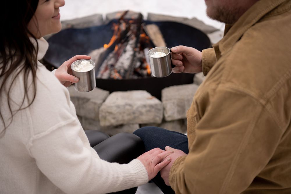 Two people hold hands while drinking from metal mugs by an outdoor fire pit.