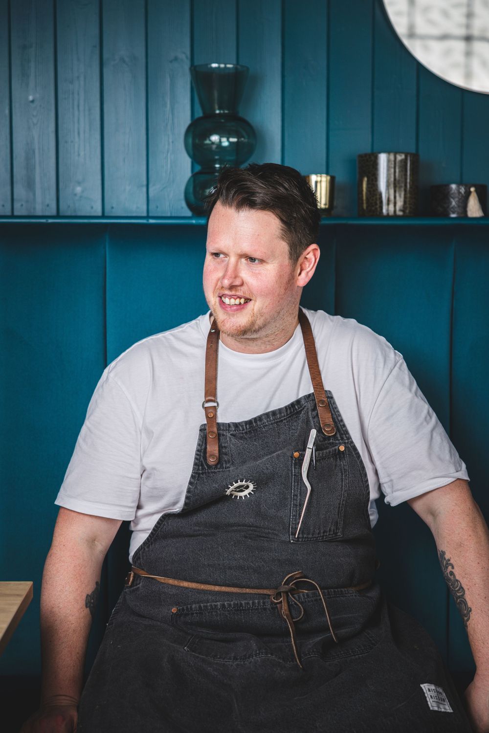 A person wearing a white t-shirt and dark denim apron sits casually in a restaurant with teal blue paneled walls.