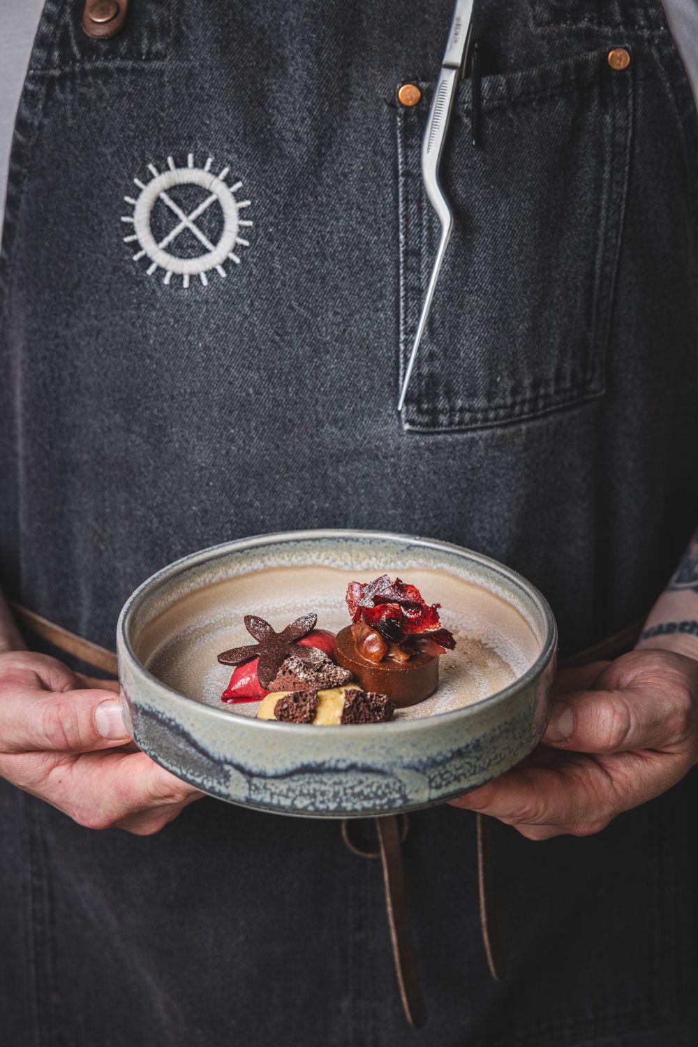 Chef holding ceramic bowl with garnished dessert, wearing denim apron with gear logo.