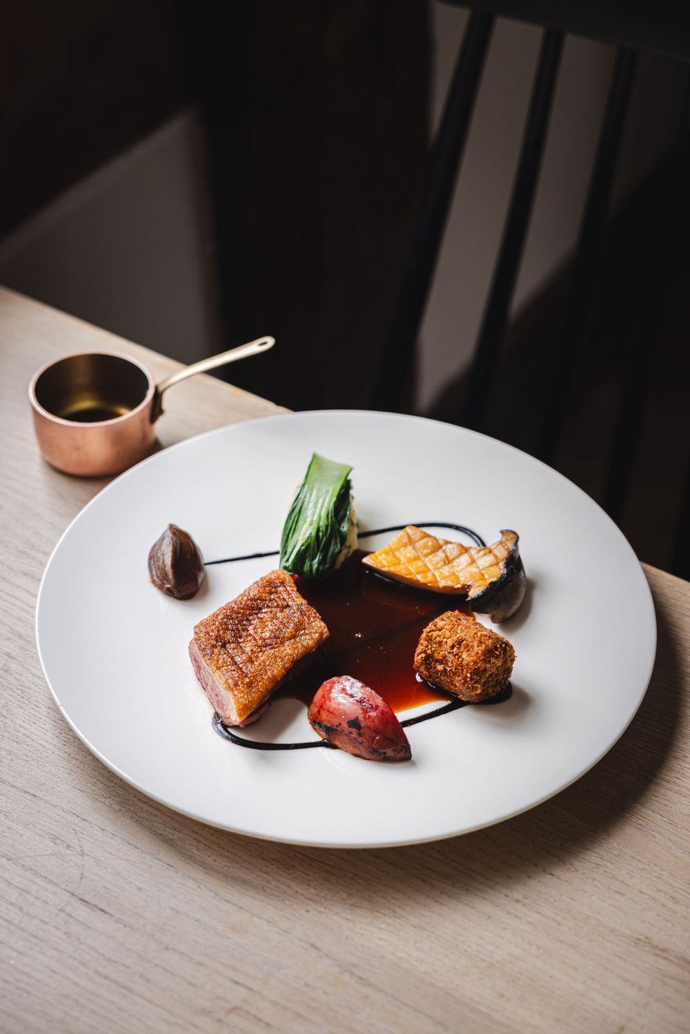 Elegant plated dish with seared protein, vegetables and sauce on white plate with copper sauce pot.