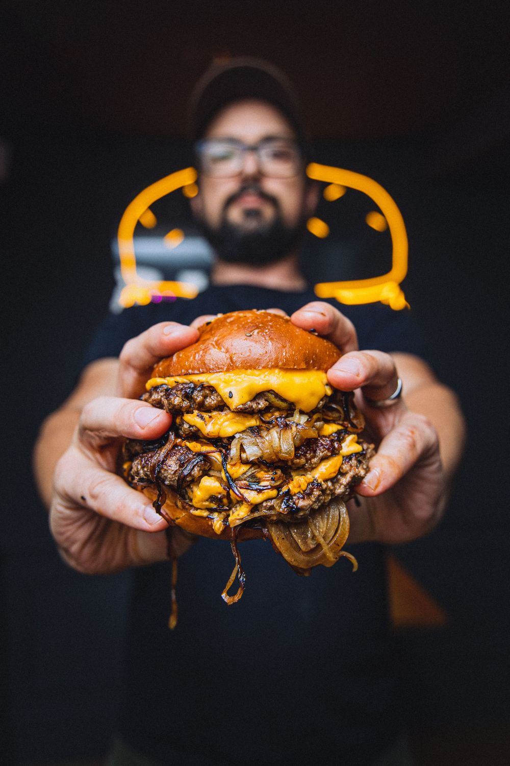 Juicy cheeseburger with melted cheese and multiple patties held in front of neon burger sign.