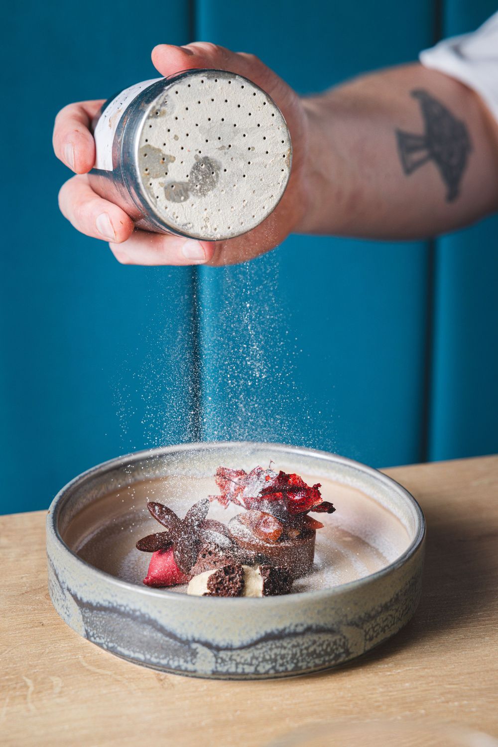 Hand sprinkling seasoning over a dish in ceramic bowl against blue background.