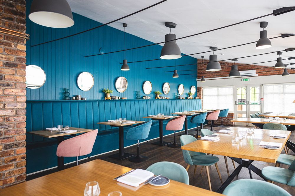 Modern restaurant interior with bright turquoise walls and pink banquette seating along a wall of circular windows.