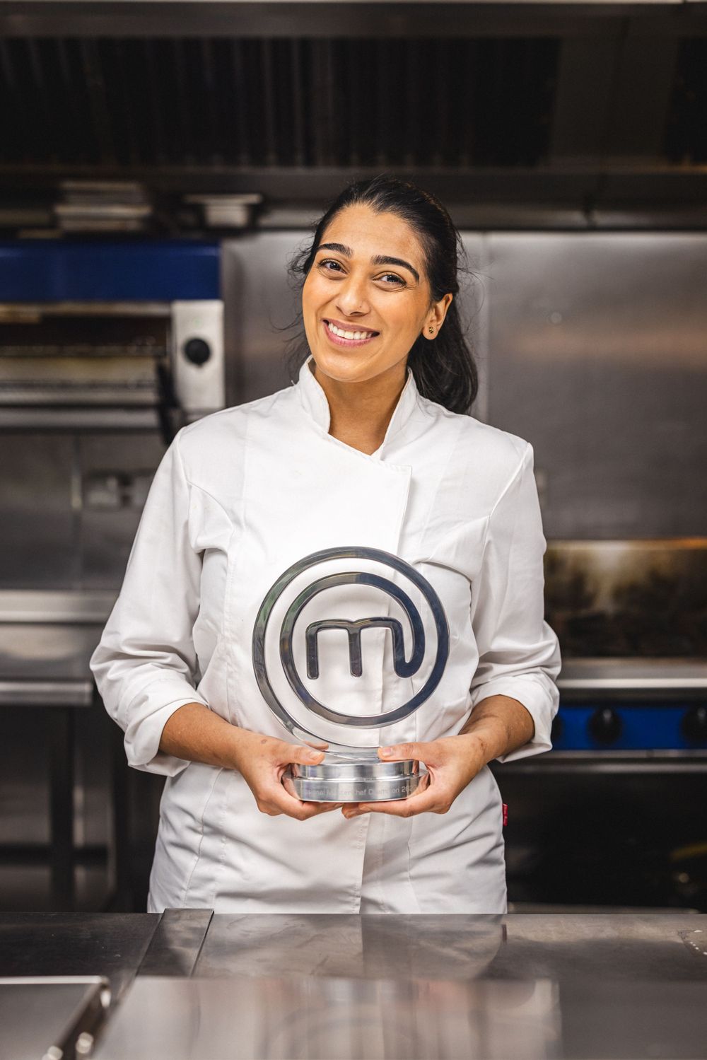 A culinary professional in white chef's coat holds a MasterChef trophy in a commercial kitchen.
