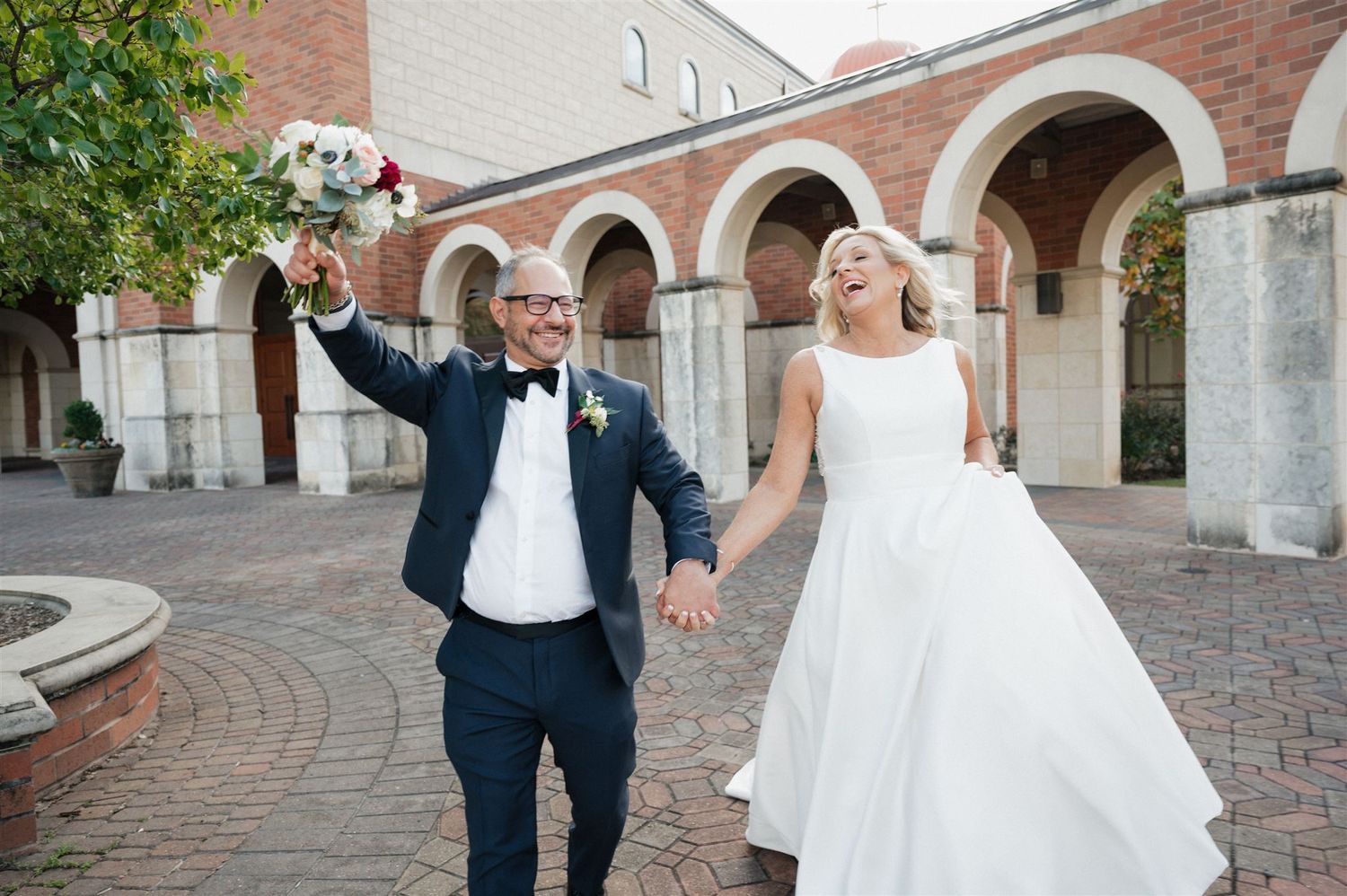 Wedding couple celebrating with raised bouquet under brick arches.