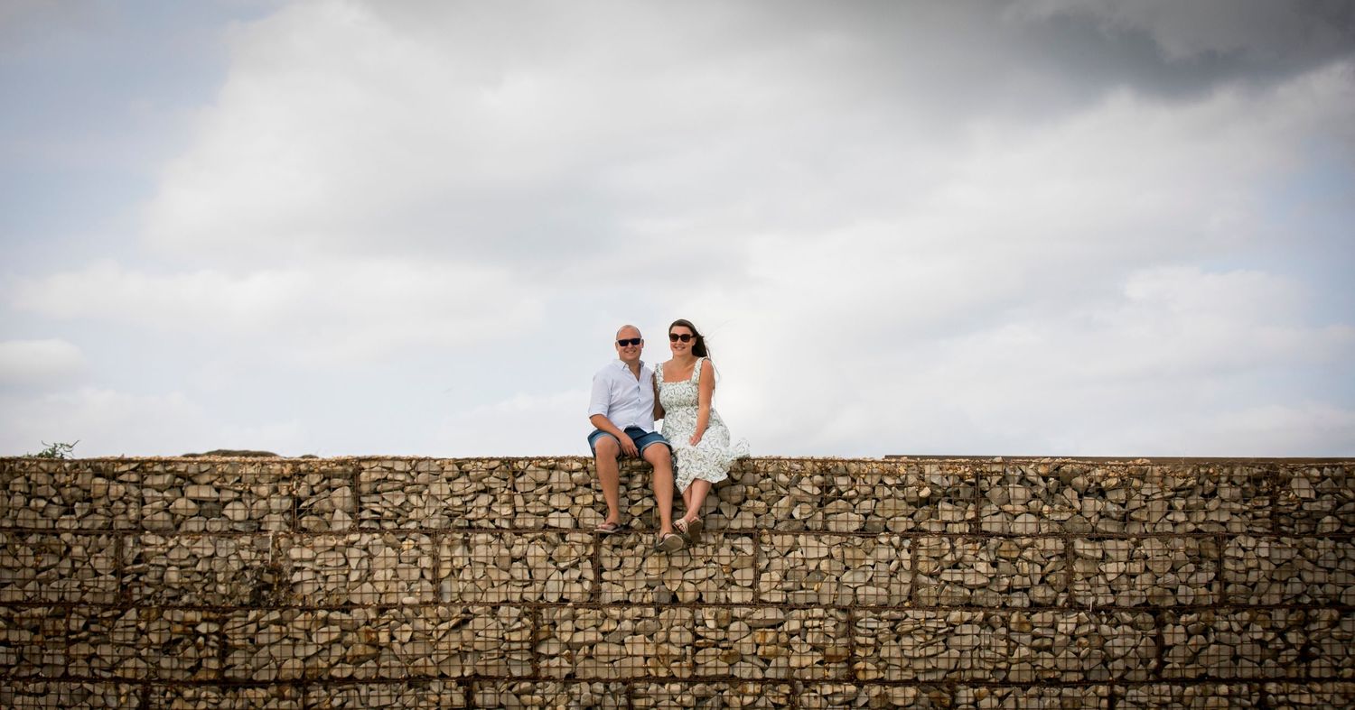 A couple shares a romantic moment on a stone wall against a cloudy sky.