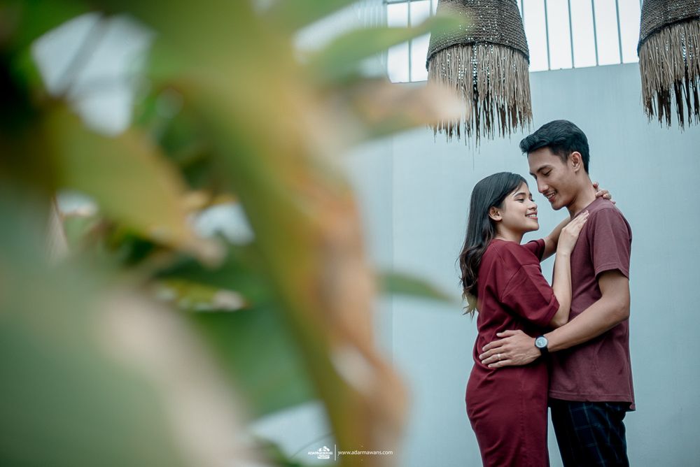 Couple in burgundy outfits embracing in intimate outdoor portrait through foliage.