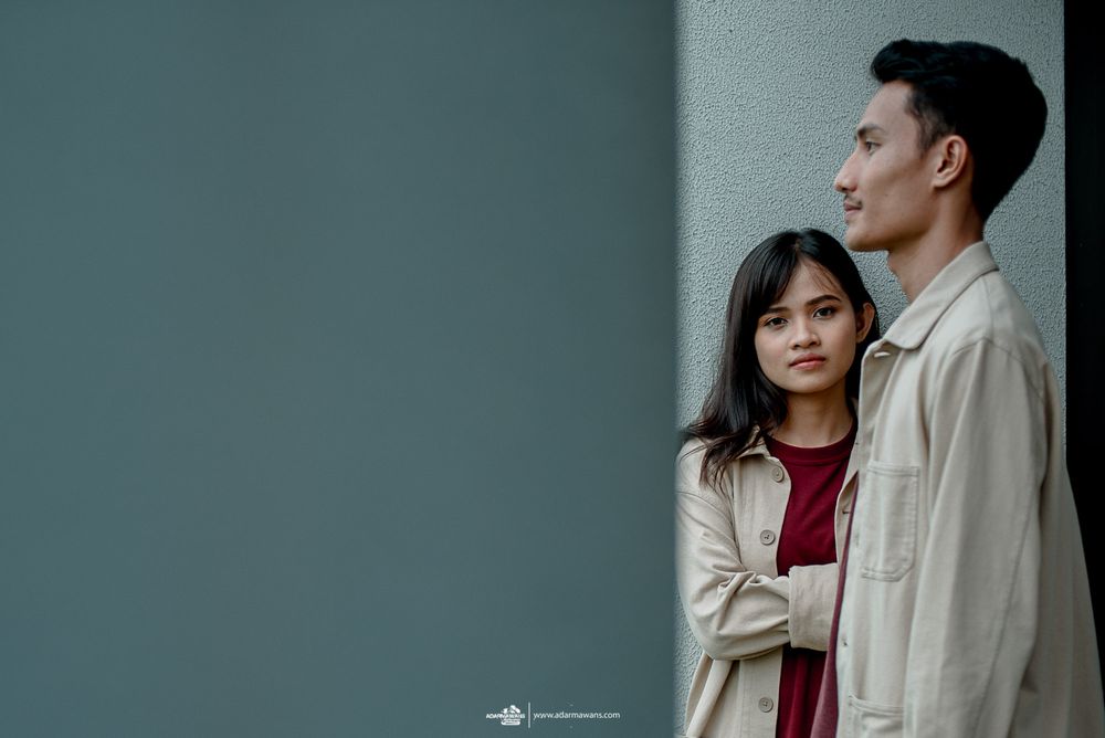 Couple in contrasting outfits pose thoughtfully against minimalist grey wall.