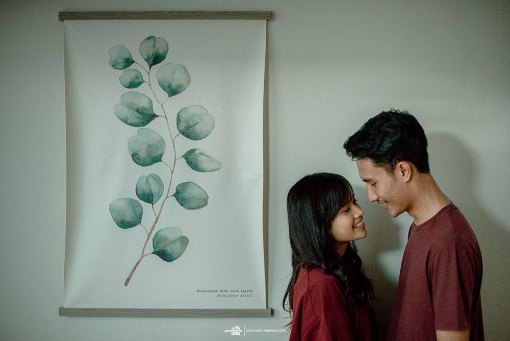 Couple in matching burgundy outfits standing against botanical wall art in minimalist setting.