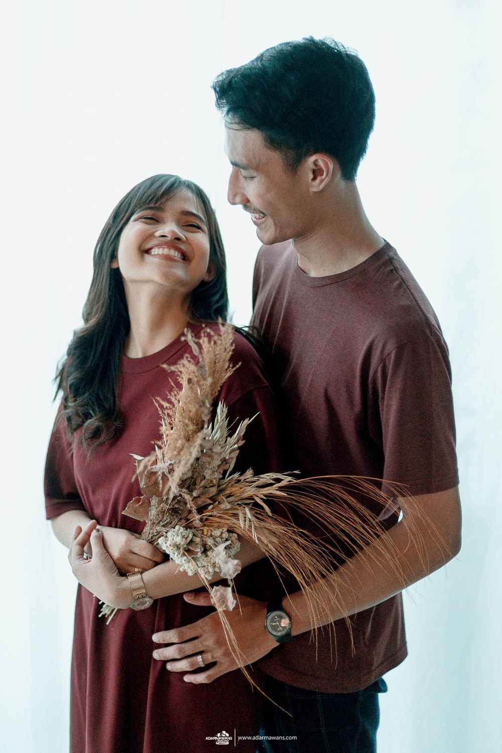 Couple in matching burgundy outfits sharing a tender moment while holding dried pampas grass bouquet.