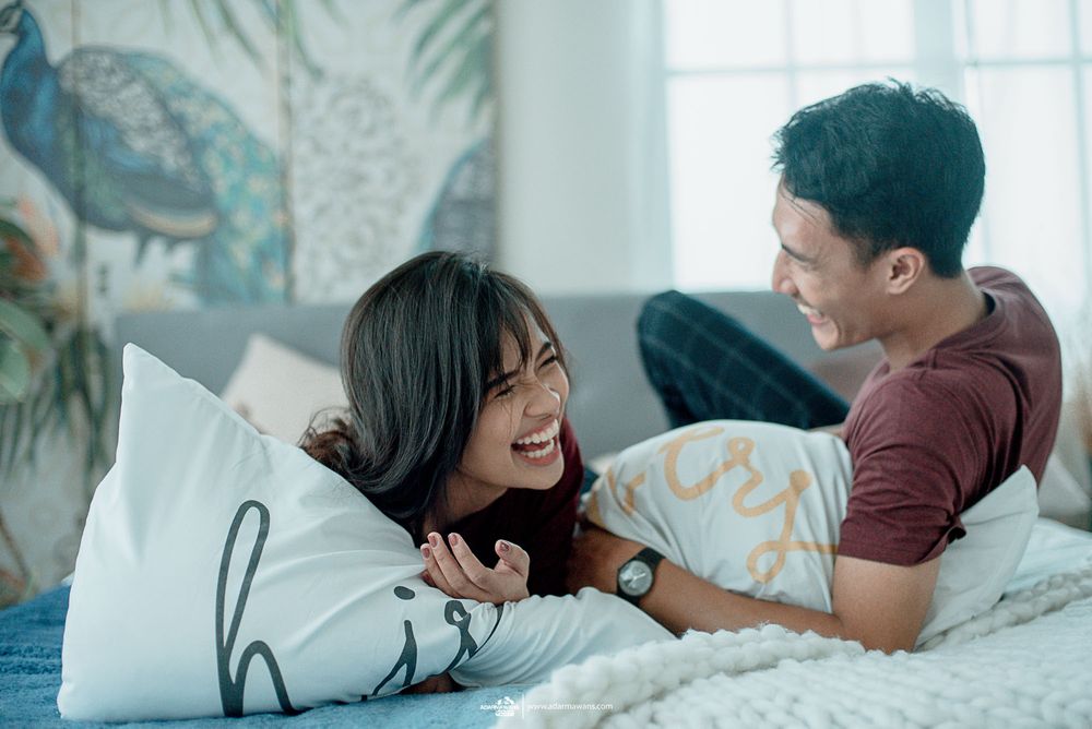 Couple in burgundy shirts laughing together on bed with pillows in bright interior.