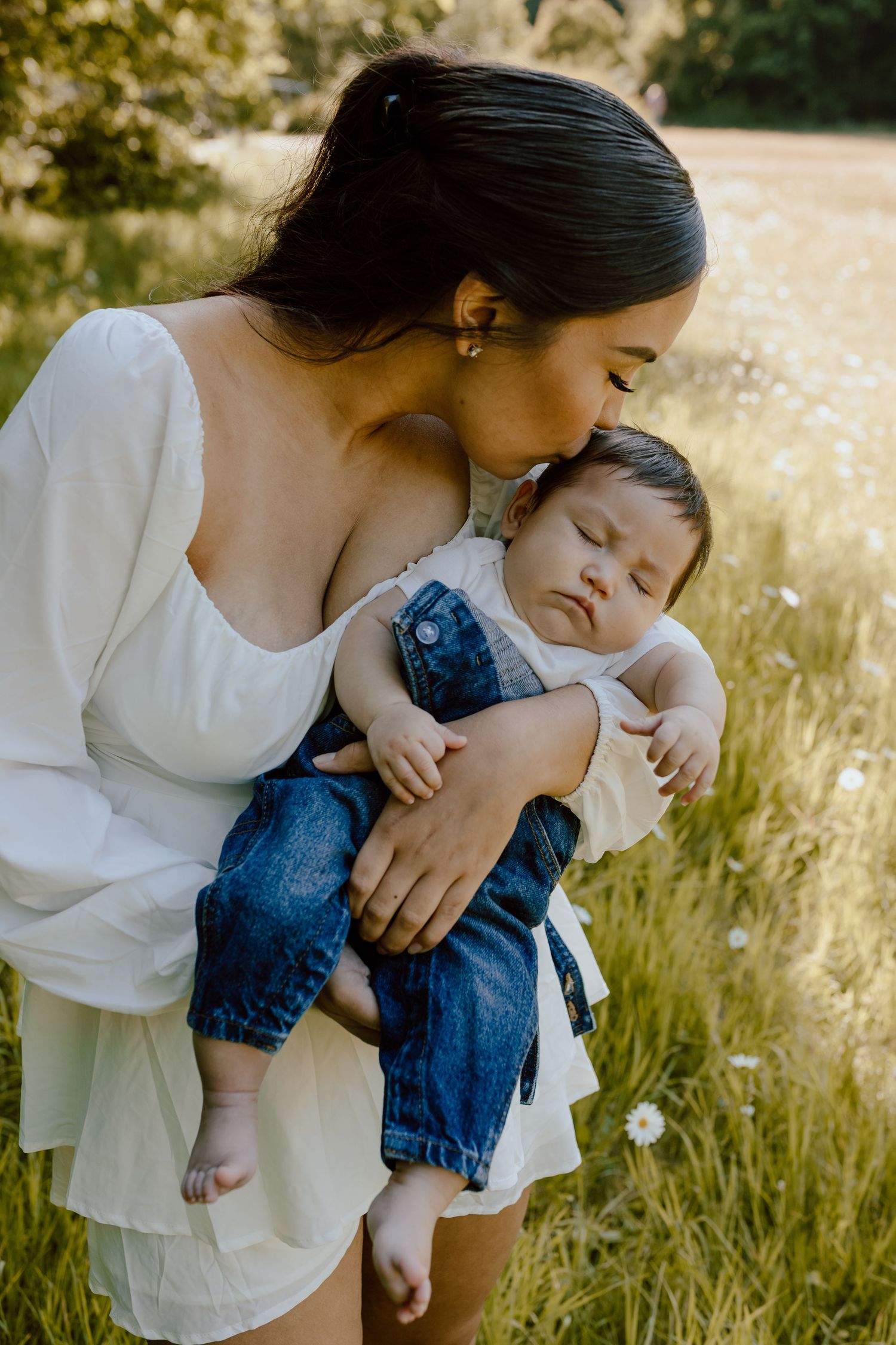 Tender moment between parent and baby in white dress amidst golden summer field.