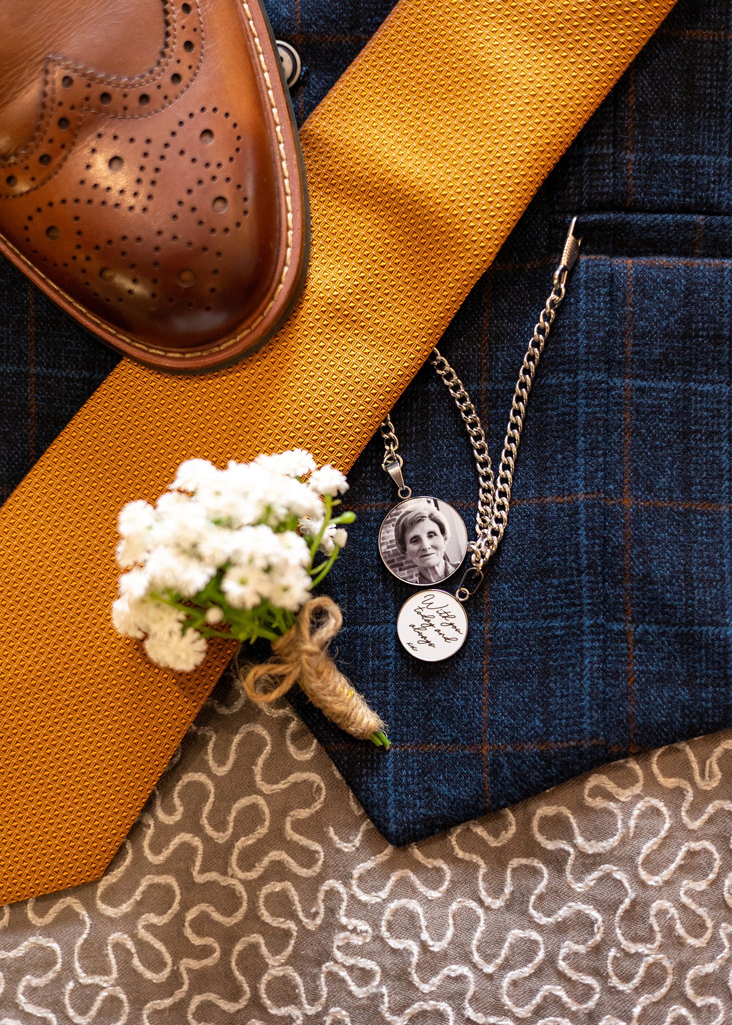 Wedding details including a brown brogue shoe, yellow tie, boutonniere and silver necklace against denim.