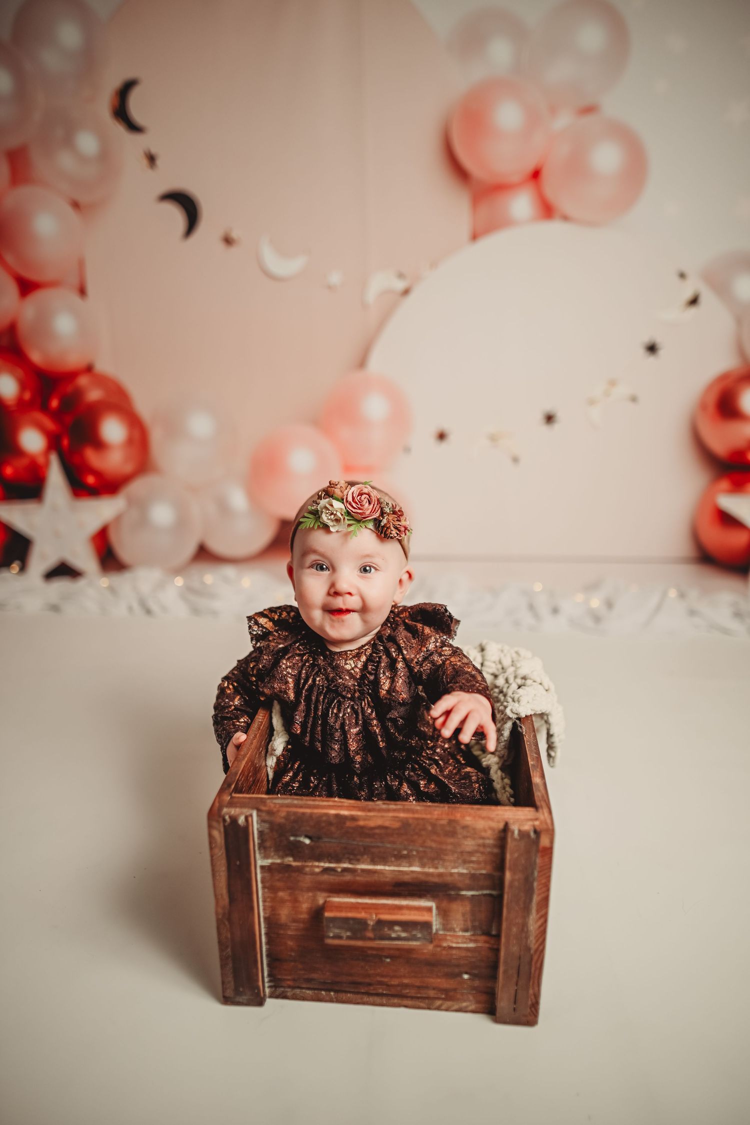 Baby sitting in wooden crate with pink balloons and crown accessory in background.