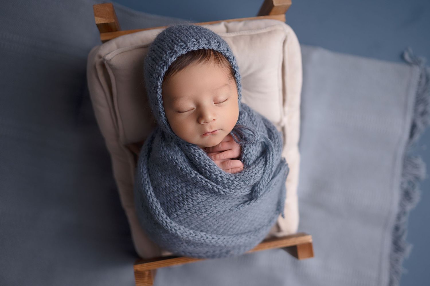A sleeping newborn wrapped in a blue knit blanket rests on a tiny wooden bed.