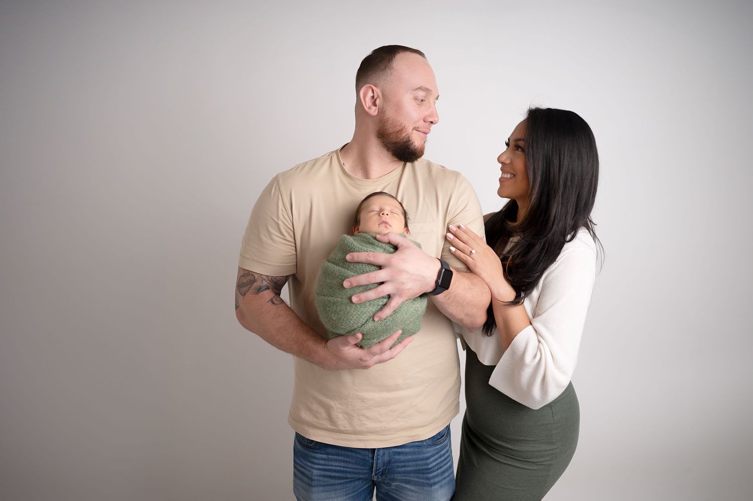 Modern studio newborn photography featuring parents in casual attire with baby in green wrap.