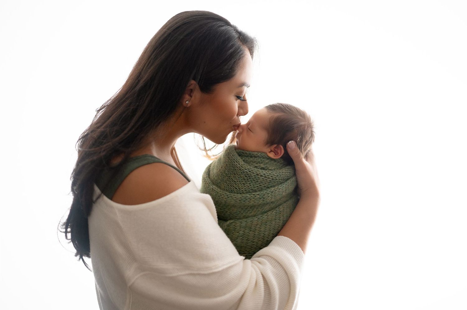 A mother in white gently kisses her newborn wrapped in green against a light background.