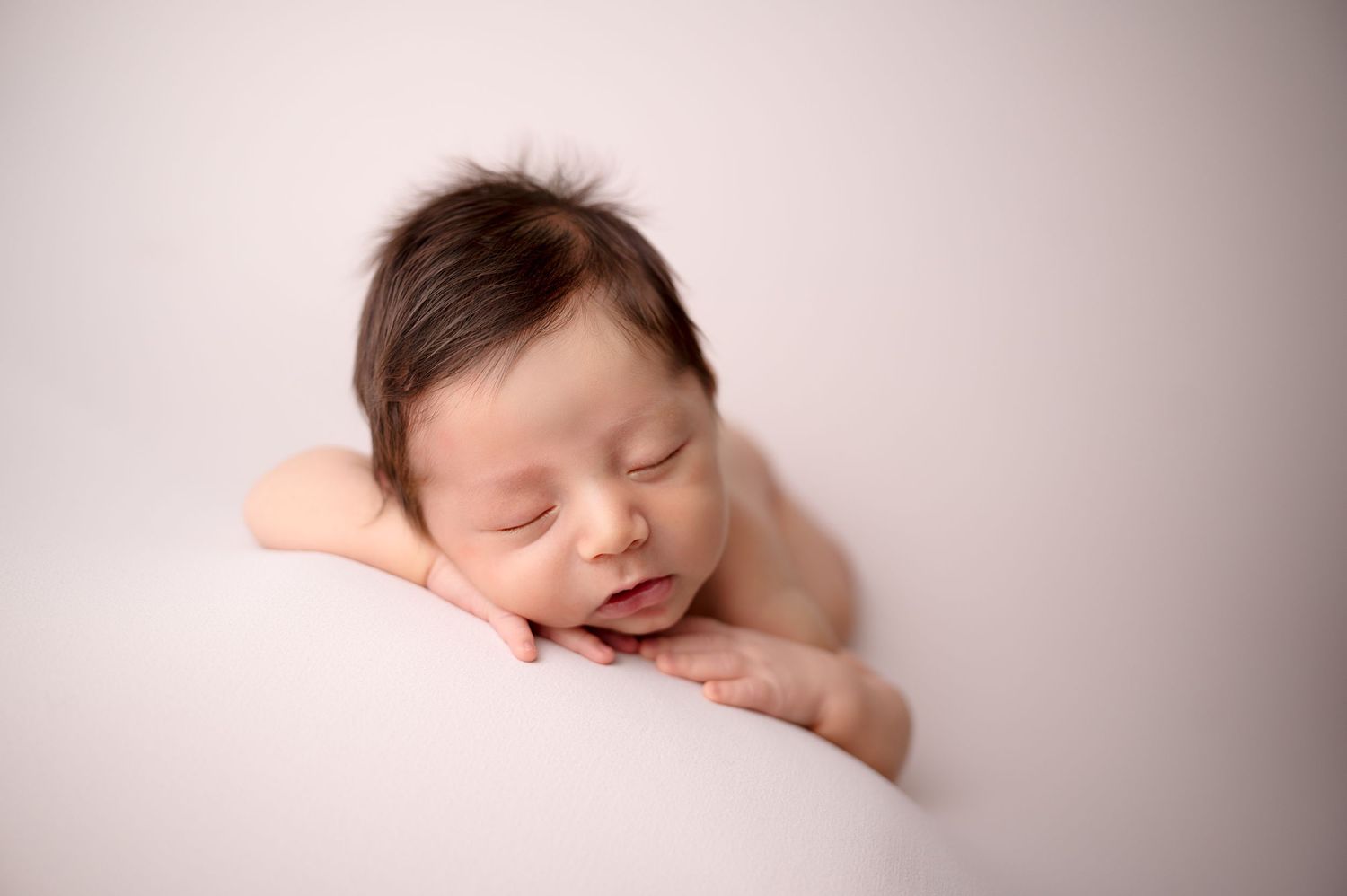 A peaceful sleeping newborn rests head on hands against soft pink background.
