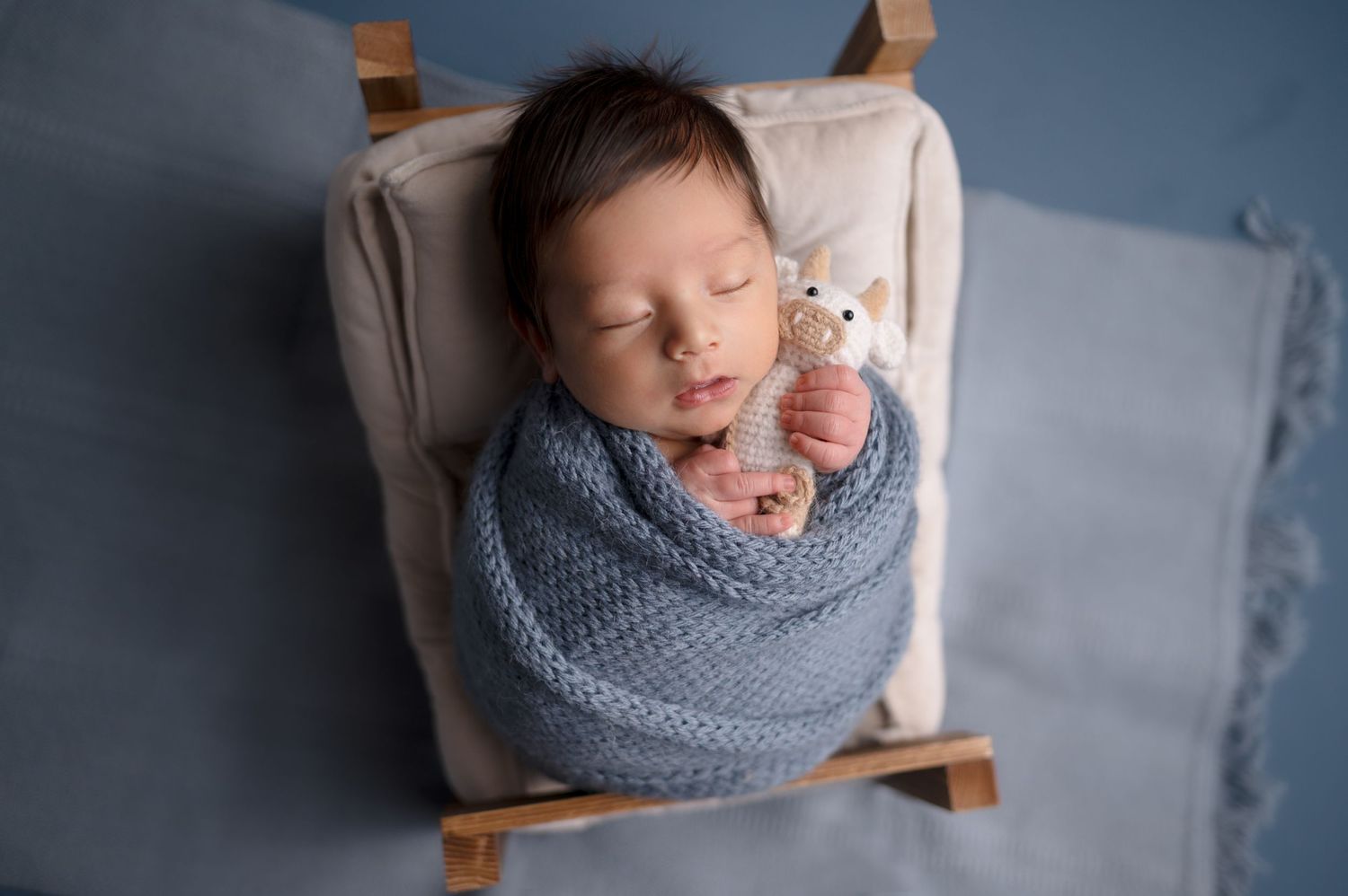 A sleeping baby wrapped in blue knit holds a tiny teddy bear on a miniature wooden bed.