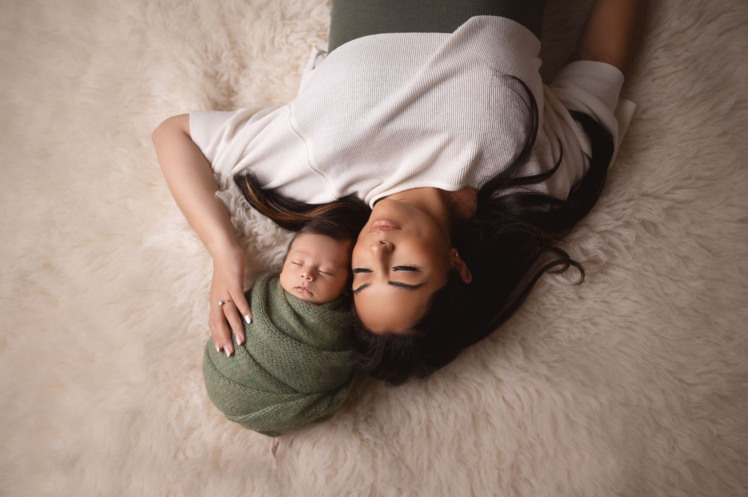 Parent and newborn lying on white fur rug in intimate overhead portrait.