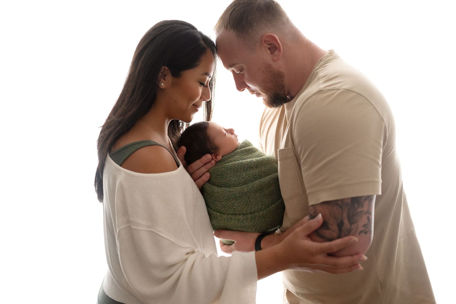 Parents sharing intimate moment with newborn wrapped in green against white background.
