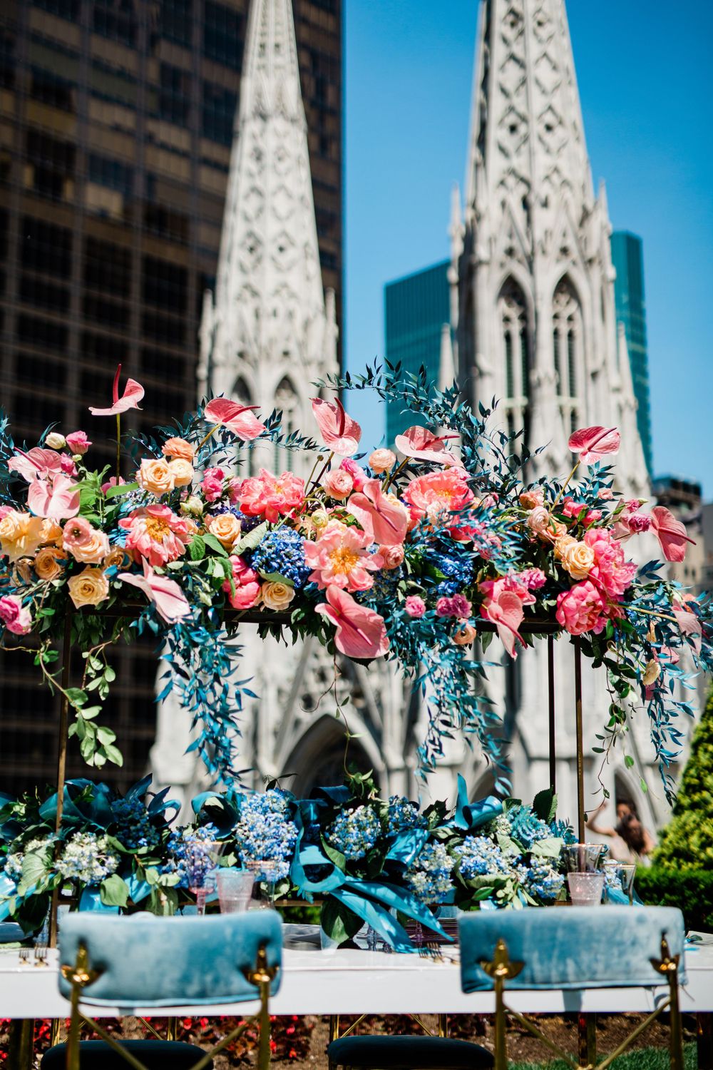 Colorful floral arrangements with pink and blue blooms displayed in front of a cathedral spire against a bright blue sky.